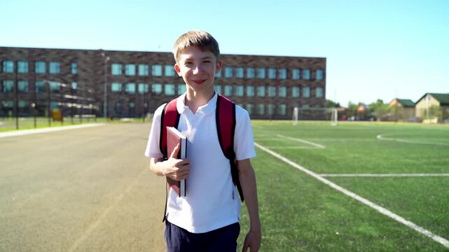 Bright sun shines on a smiling boy. He stands near a school with green field. He carries books, wearing a backpack. He looks directly at the camera with joy