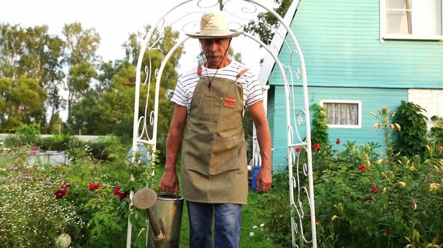Man stands in garden, holding watering can. He wears hat and apron, smiling slightly. White arched trellis frames the scene behind him. Vibrant flowers surround him, blooming in soft light