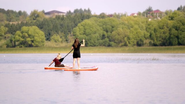 Two people paddle on calm waters. One stands, one sits on a red paddleboard. They glide past green trees and distant houses. The sun shines softly on their calm expressions
