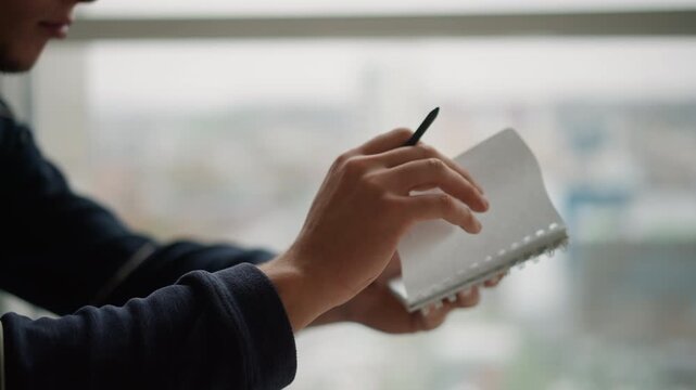 Close up of person writing in notebook with pen focusing on hands and stationary object interaction. Notebook pages are visible during action. Soft natural light fills indoor setting
