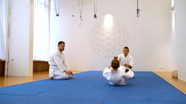 Young boy practices judo with instructor in bright studio. He falls backward while another observes calmly. Blue mat contrasts with white walls and wooden floor. Scene shows discipline, focus