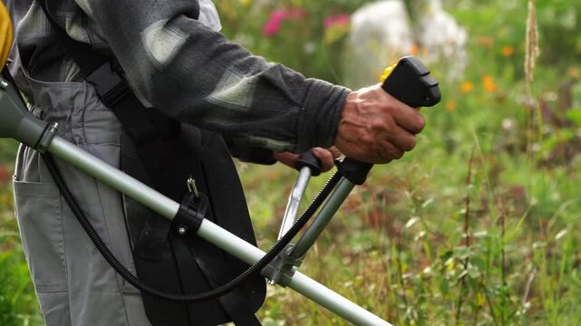 Worker grips tool, moves through tall grass. Yellow engine powers cutting tool. Green field dotted with wildflowers. No animals or plants identified