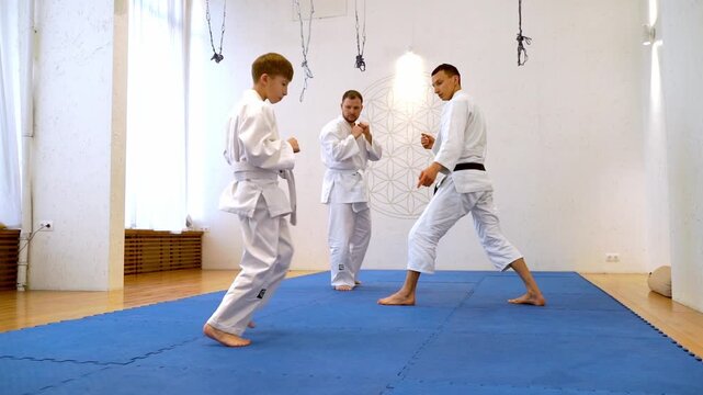 Two children train karate on blue mat. One child kneels, another lies on back. Adult watches from seated position. Room has white walls and hanging ropes. Scene shows discipline and focus