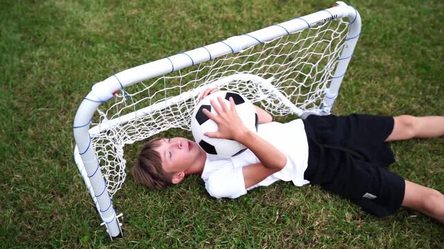 Young boy lies on grass, holding soccer ball. He rests under small goal net, smiling. Child wears white shirt, dark shorts. Joyful play outdoors, on green field. Perfect for sports, childhood, fun