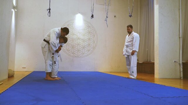 Two men in white uniforms bow respectfully. One kneels, eyes closed, showing deep focus. Another stands, observing with calm patience. Bright light shines behind them, illuminating the blue mat