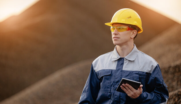 Industrial worker with tablet and yellow safety helmet in sand quarry at sunset for site inspection