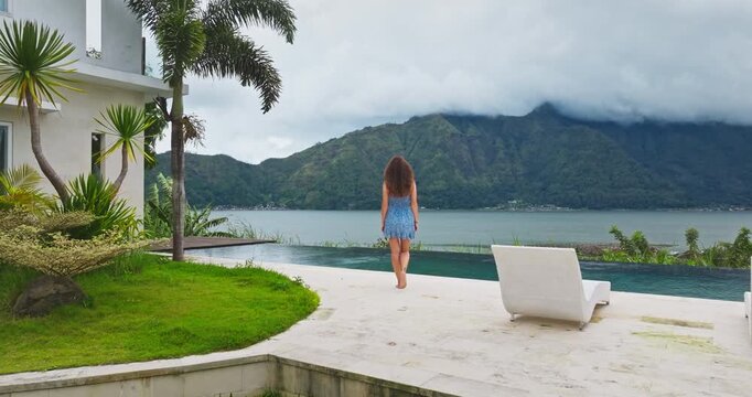 Woman in a blue dress walking barefoot towards an infinity swimming pool, enjoying the luxurious tropical villa surroundings with a stunning view of Mount Batur and Lake Batur in Bali, Indonesia