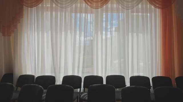 Back view of empty chairs in an empty conference hall. The wind gently moves the thin white curtains, showing the sky outside the window