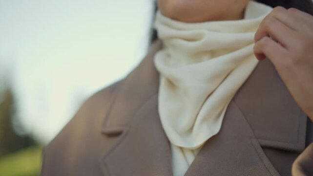 Close up of woman adjusting her cream colored scarf around neck while wearing brown coat soft lighting and calm expression. Hand movement is visible as woman positions fabric with long dark