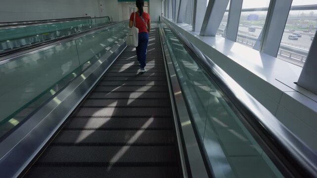 Woman walks along a moving walkway inside a modern airport terminal, carrying a tote bag beside glass railings and large windows