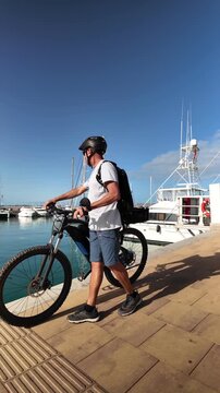 Slow motion shot of an active mature man with a backpack walking with his electric bicycle along a pier in a marina on a sunny day, promoting a healthy lifestyle and sustainable commuting