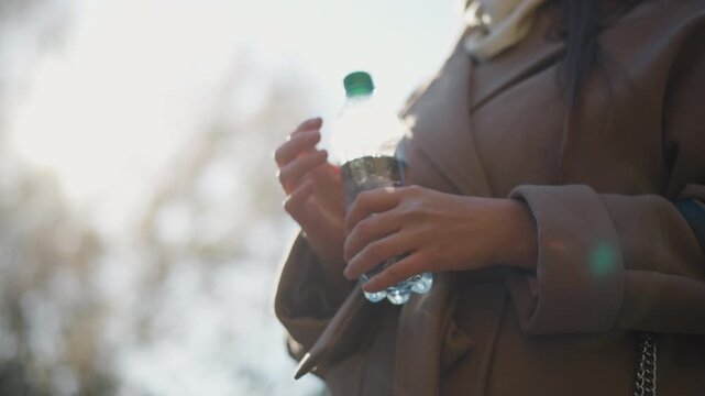 Close up of woman holding and slightly adjusting her water bottle while wearing brown coat with soft sunlight creating lens flares. scene focuses on hands and lower torso showing interaction