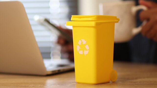 Yellow recycling bin sitting on office desk with a businessman using a laptop and drinking coffee, promoting sustainability and waste management in the workplace