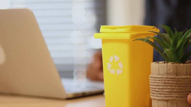 Office worker typing on a laptop with a yellow recycling bin on the desk, illustrating a sustainable business practice and environmental awareness in the workplace