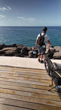 Mature man stopping during a ride with his electric bicycle on a seaside promenade, looking at the calm blue sea on a sunny day. He is practicing a healthy and sustainable lifestyle