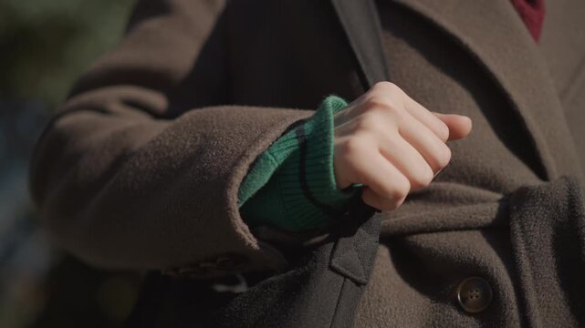 Close up of young girl hand gripping strap of her black bag wearing green ribbed sleeve and brown coat. scene shows consistent soft lighting on textured fabric details like woolen material
