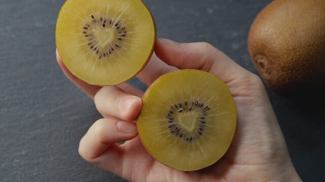 Macro shot of female hand holding two halves of ripe golden kiwi on dark gray background. Fingers gently rotate one half, showing rich juicy texture for healthy eating, gourmet food, culinary content.