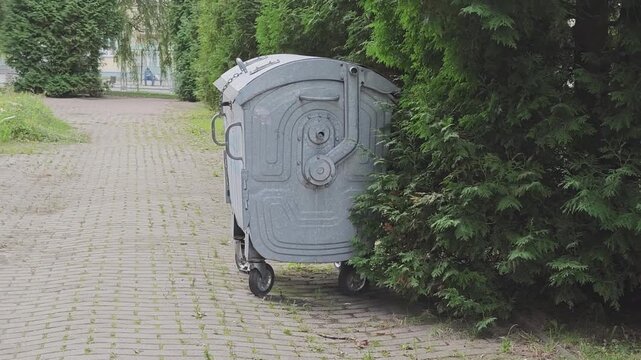Large gray metal dumpster on wheels standing on a paved pathway next to a green hedge. Outdoor garbage collection and waste management concept
