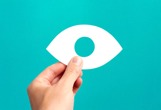 A persons hand holding a white paper cutout of an eye shape with a circular pupil against a tealcolored background symbolizing vision