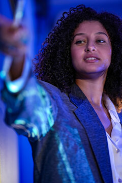 Businesswoman with curly hair explains the glowing data visualization rim lit in gold while speaking beside a deep blue wall at a high tech business conference about artificial intelligence