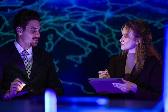 Businessman and young female colleague react to a figure on the screen seated like news anchors before world map and interface graphics while the team records a technology explainer in the