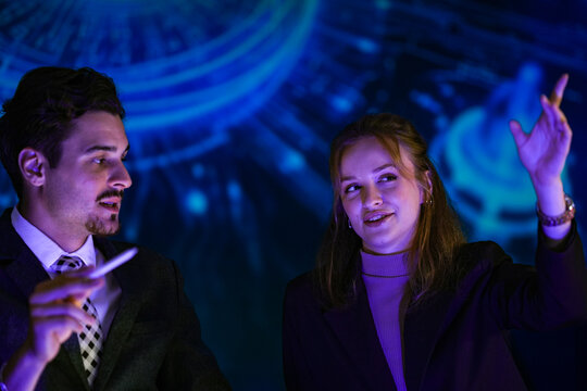 Businessman and young female colleague standing and debating beside globe and brain projections raise a stylus to highlight a detail while the team records a technology explainer in the
