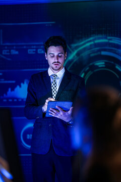 Businessman in dark suit and tie presenting with a tablet as silhouetted viewers watch reviews notes on a digital tablet while the team records a technology explainer in the darkened studio