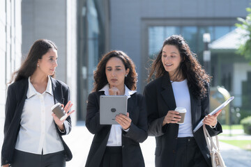 Three female colleagues in matching black business suits, talking as they walk in step, move as a group across a sunlit forecourt with coffee cups and handbags in hand. © ultramansk