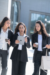 Three female colleagues in matching black business suits, talking as they walk in step, move as a group across a sunlit forecourt with documents tucked under one arm, shot outdoors. © ultramansk