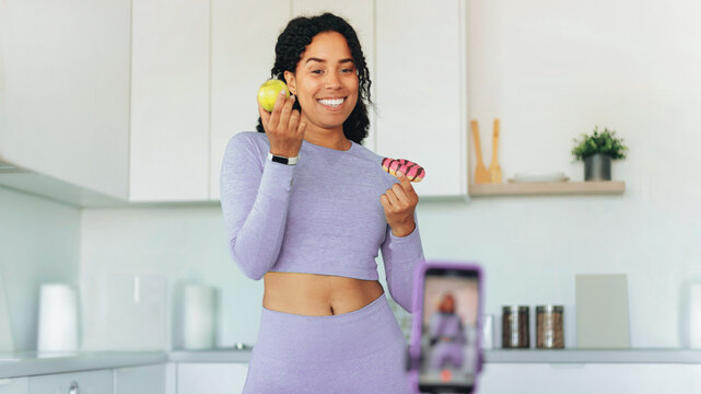 Smiling African American Woman Holding An Apple And A Doughnut In A Modern Kitchen, Recording A Video. Cheerful Female Influencer Promoting Healthy Eating And Lifestyle With Visual Engagement.