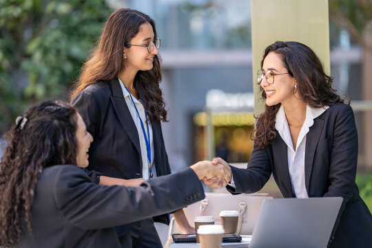 A diverse team of three Hispanic businesswomen in dark suits, smiling with mutual respect, welcome a new partner to the table reaching across the table to shake hands.