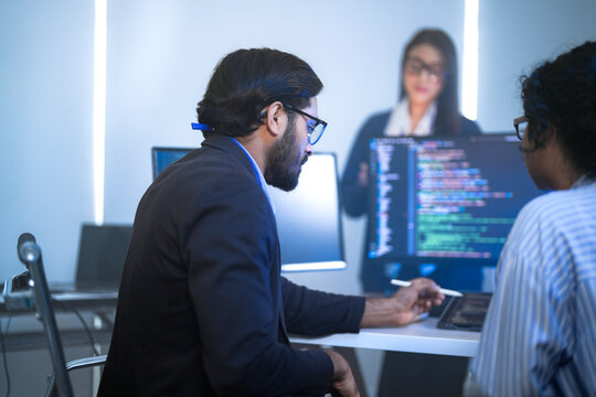 A male software engineer in a suit checking project data on a tablet device, at a long desk with code monitors against a bright window; side profile view of software development.