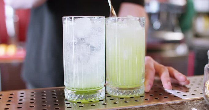 Stirring drink. Cocktail bar. Bartender hand mixing green liquid with ice cube sparkling water long spoon in glass on metal counter.