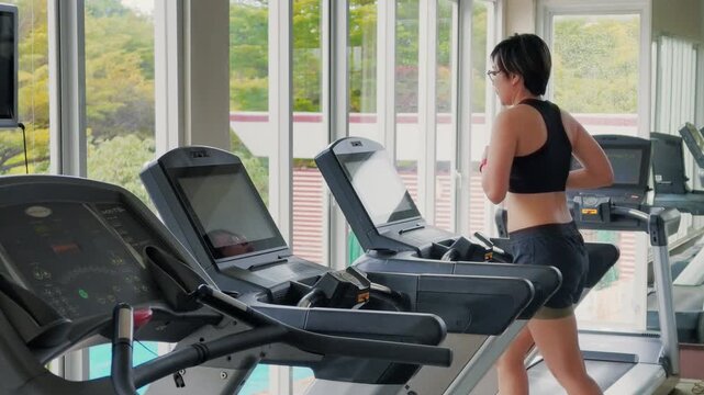 Wide shot of an active Asian woman in her forties running on a treadmill in a bright modern gym. She wears a black sports bra, enjoying daily cardio exercise by large windows with natural daylight.