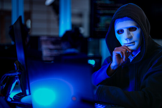 Profile of a mysterious anonymous hacker in a black hoodie and white mask infiltrating a corporate network infrastructure under blue lighting.