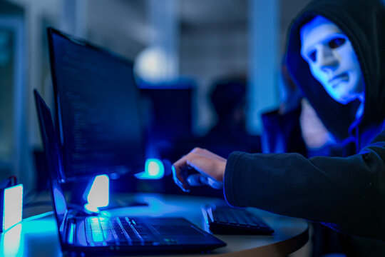 Profile of a mysterious anonymous hacker in a black hoodie and white mask infiltrating a corporate network infrastructure under blue lighting.