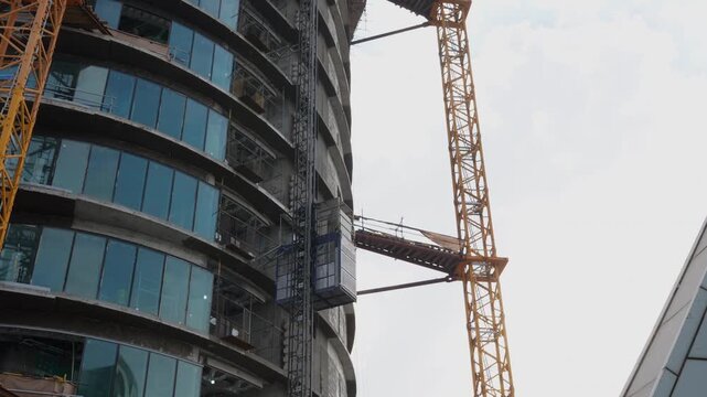 Construction elevator rises beside a tower crane on a high rise building under construction with a curved glass facade