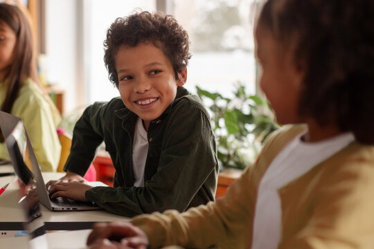 Smiling mixed-race elementary school boy looking up from his laptop at classmate