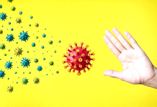 Human hand raises to stop approaching aggressive red coronavirus particle and swarm of smaller blue and green virus particles against a bright yellow background