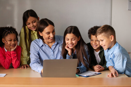 Excited female teacher and five diverse elementary school students gathered around laptop