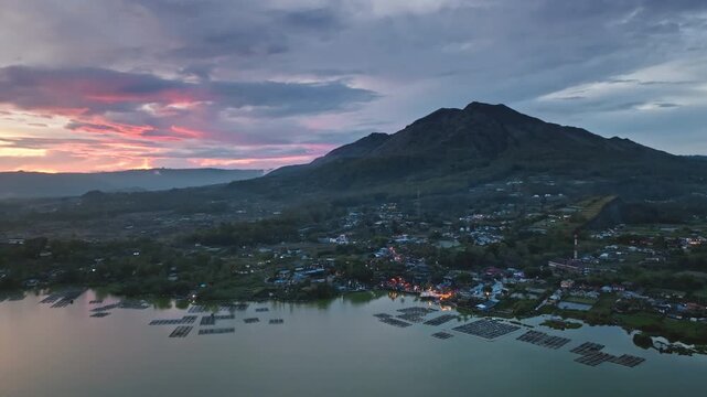 Aerial view mount Batur volcano and lake Batur with fishing cages, small villages with glowing lights, and dramatic twilight sky during a scenic sunset in Kintamani, Bali, Indonesia. Drone flight