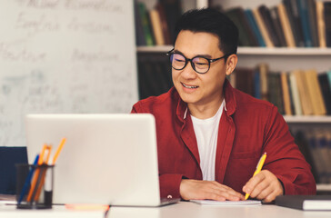 Obraz premium Smiling Asian Man Working At Laptop In Office Setting, Taking Notes With Pencil, Focused On Learning. Young Adult Engaged In Education And Skill Development With Positive Attitude.