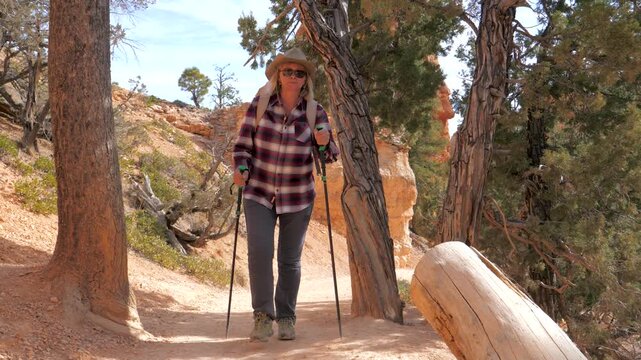 Fit mature woman with trekking poles and backpack hiking along dirt trail in canyon, surrounded by red rock formations and pine trees, enjoying an active outdoor lifestyle on a sunny day