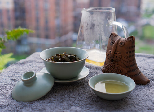 Traditional Chinese Gongfu tea ceremony set with an open green celadon porcelain gaiwan showing brewed oolong leaves, matching teacup, glass chahai pitcher, and clay cat statuette. Tableware arranged.