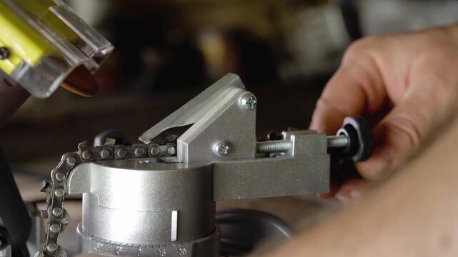 Man adjusts small chain sharpening machine with focused hands in workshop, turning knob and guiding metal chain under yellow tool. Industrial close up shows careful repair work and precise