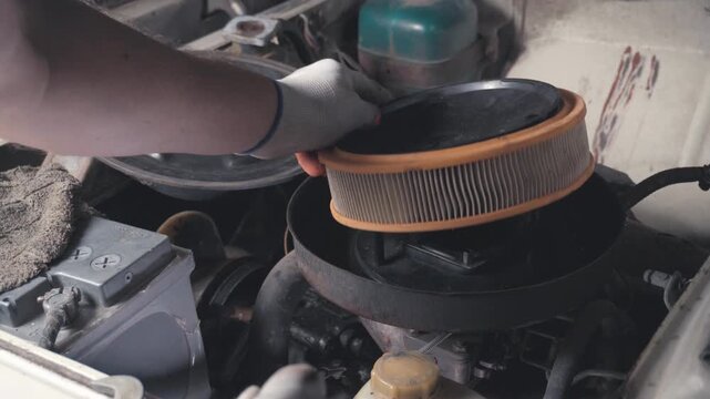 Gloved auto mechanic handles round air filter during engine service, moving carefully over exposed parts. Mechanic opens housing, swaps filter element, and works in gritty industrial style