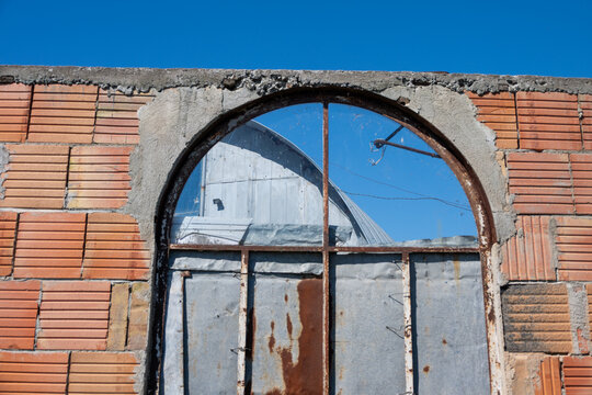 an unfinished terracotta brick wall featuring a rusty, arched window frame partially boarded with scrap metal,
