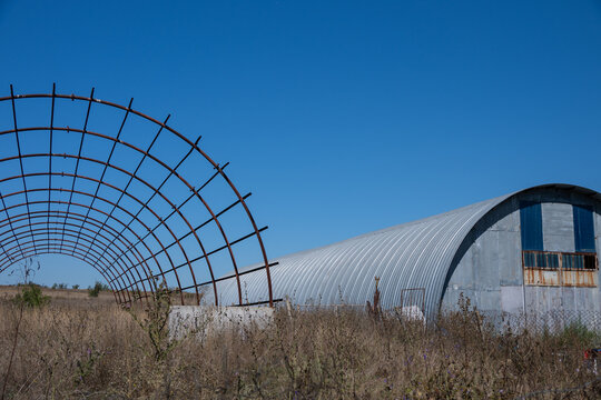A large corrugated metal Quonset hut stands alongside an exposed steel rib framework in an open, dry rural field under a clear blue sky.