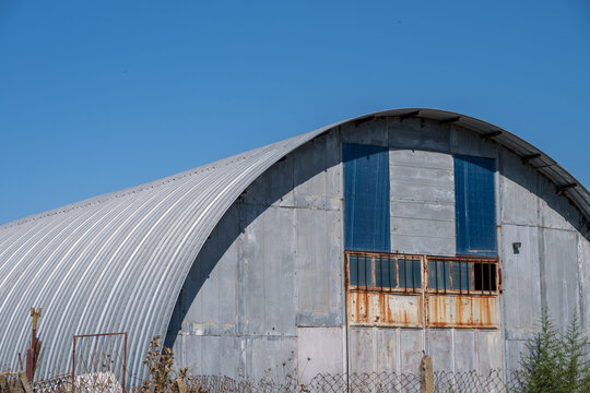 a large, weathered corrugated metal Quonset hut standing behind a wire fence in an overgrown field of dry grass under a clear blue sky.