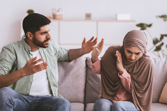 Worried Arab Woman Wearing Hijab Trying To Avoid Conflict With Angry Arab Man, Arguing While Sitting On Sofa At Home. Emotionally Charged Atmosphere Indicating Relationship Tension.